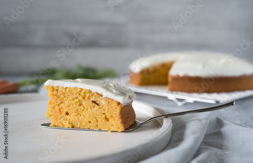Stück frisch, selbstgebackener Karottenkuchen mit frischkäsetopping auf weißem Teller serviert vor hellem Hintergrund bei Tageslicht