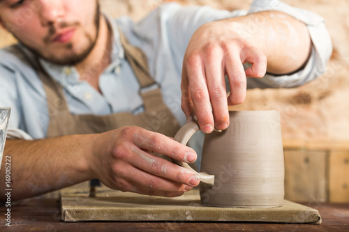 potter, stoneware, ceramics art concept - craftsman's hands connect the cup and the handle made of raw clay, master fingers work with pieces of clay, male sits at a workshop behind the table
