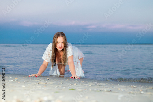 Fotografija beautiful sexy girl in a white bathing suit and tunic sits on the shore of a deserted beach