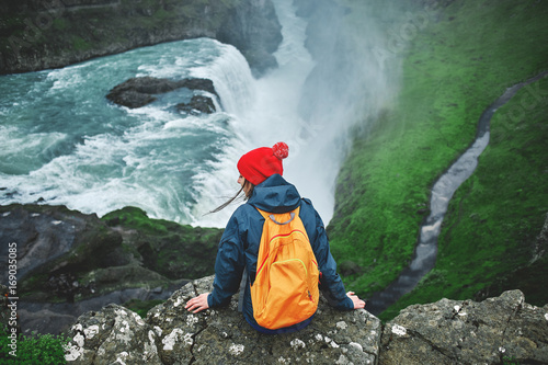 Girl in warm clothing, in red knitted hat and small orange backpack sits on the cliff on background of Gullfoss waterfall in Iceland. View from above photo