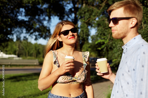 Beautiful young couple in stylish sunglasses resting in park  enjoying coffee-to-go. Outdoor shot of blonde girl admiring ideas of her smart creative husband. Selective focus on woman s face