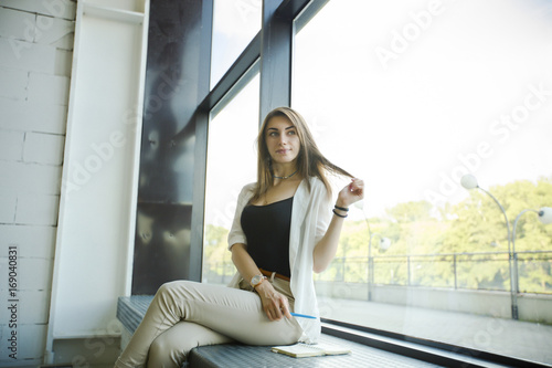 Young beautiful trendy woman sitting on the windowsill in office and smiling. 