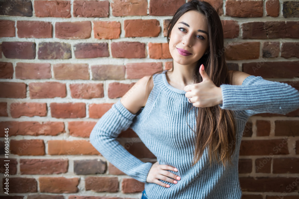 Beautiful young woman in casual wear with thumbs up, smiling, standing against white brick wall