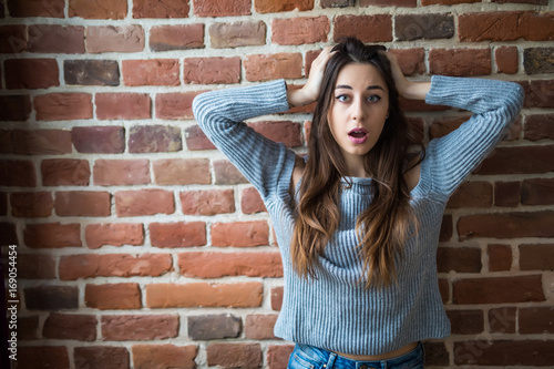 Beautiful young woman in casual wear surprised look and standing against white brick wall
