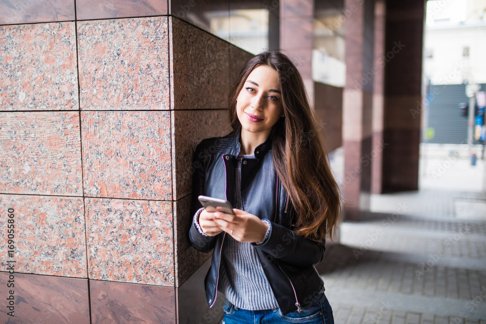 beautiful young woman is using an app in her smartphone device to send a text message in front of a building background