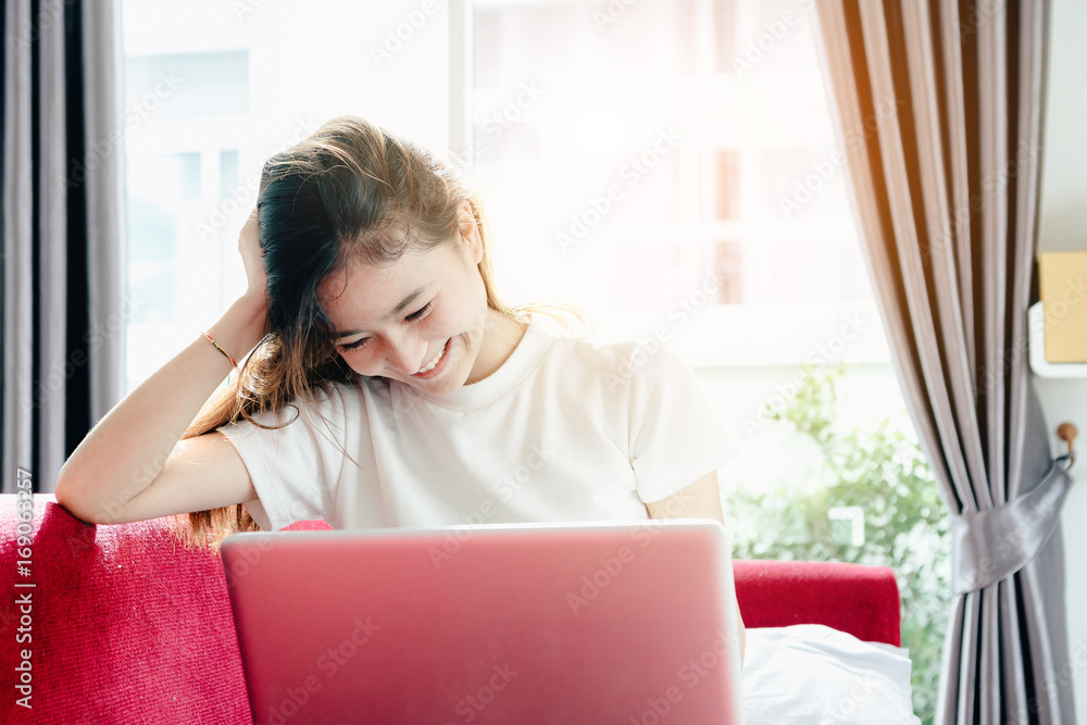 Asian teenage girl using a laptop computer to check her orders online and drinking coffee on a red sofa in her home. Asian teenage girl using a laptop computer to check her orders online and drinking coffee on a red sofa in her home.