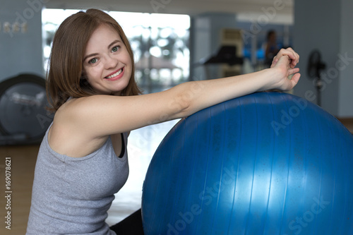 Portrait of a slim smiling girl with green eyes sitting near blue gymnastic ball looking happy and fit with training apparatus on background