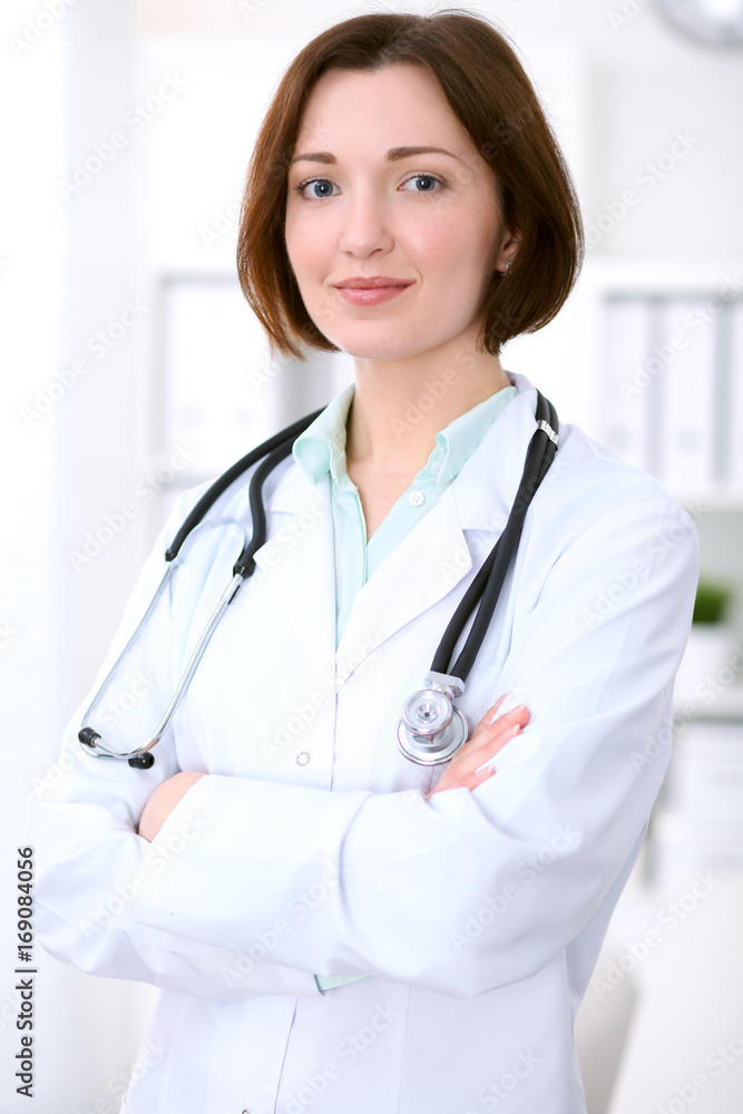 Young brunette female doctor standing with arms crossed and smiling at hospital Young brunette female doctor standing with arms crossed and smiling at hospital