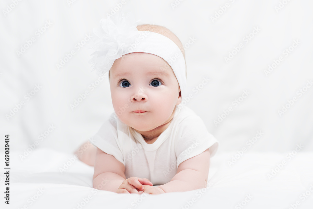 Cute baby girl in white shirt, with bow flower on head, lying on belly on soft blanket, indoors Cute baby girl in white shirt, with bow flower on head, lying on belly on soft blanket, indoors
