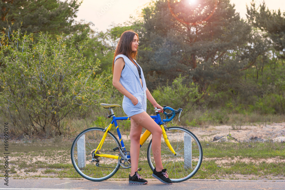 Woman stands with a bicycle on a road in the park in the sunset rays of the sun