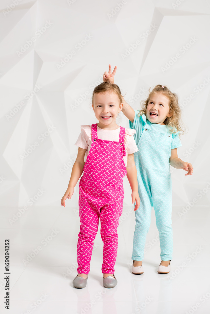 Two little girlfriends in the identical overalls of different colors in a studio with white walls Two little girlfriends in the identical overalls of different colors in a studio with white walls