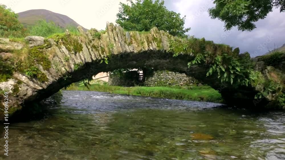 Reveal from an old stone footbridge over a flowing river in the Lake District, UK. Reveal from an old stone footbridge over a flowing river in the Lake District, UK.