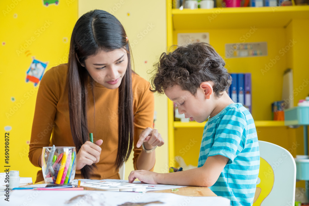 © weedezign - Asian woman teacher teaching boy kid to paint color book on table in classroom,kindergarten education school