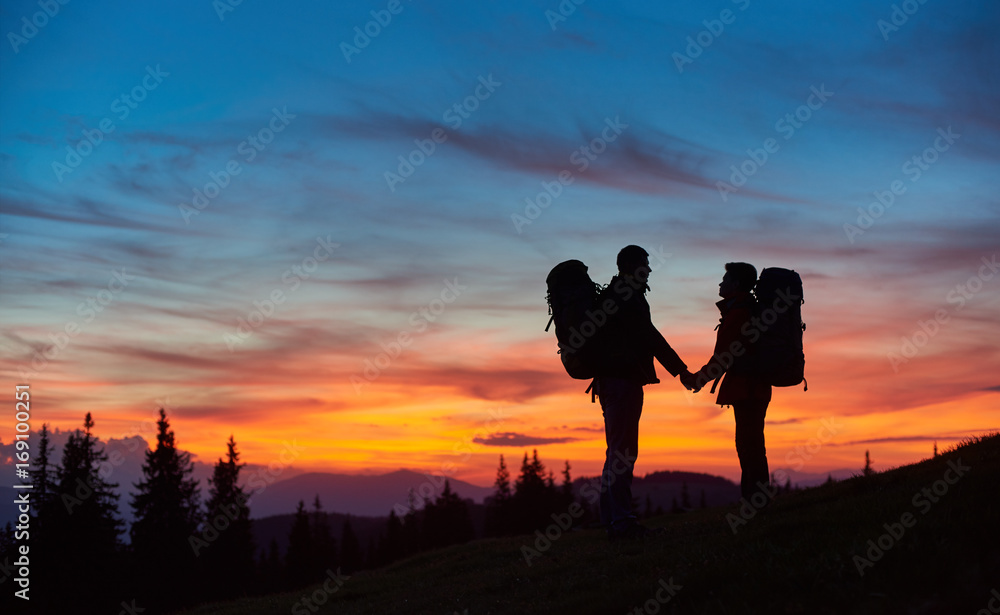 Loving couple tourists hiking together holding hands standing with their backpacks on top of a mountain during stunning sunset people love silhouettes meadow active relationships