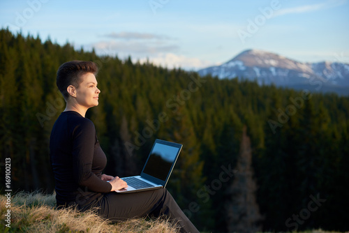 Happy young attractive woman working on her laptop outdoors enjoying view of the mountains copyspace