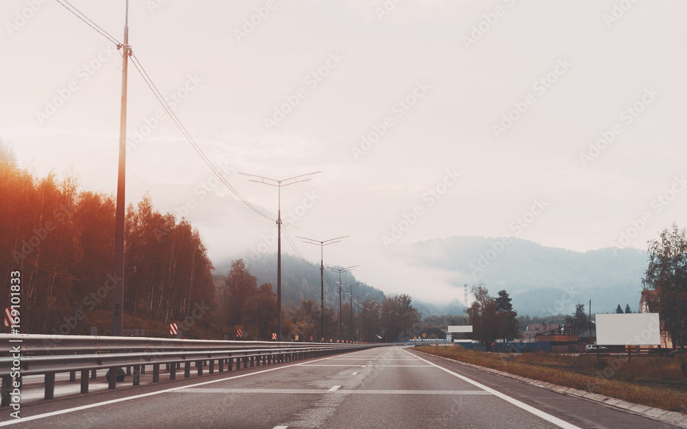 View of empty highway on sunny misty morning with mountains in distance partly covered by low clouds, mock-up of road banner on the right, Altai district, Russia