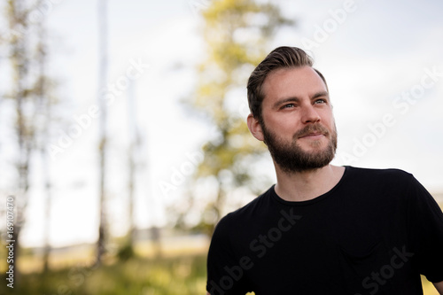 Laid back bearded man wearing a black shirt, standing outdoors on a sunny summer day looking away from camera with a smile on his face.