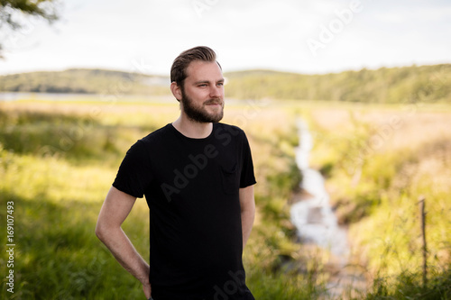 Laid back bearded man wearing a black shirt, standing outdoors on a sunny summer day looking away from camera with a smile on his face.