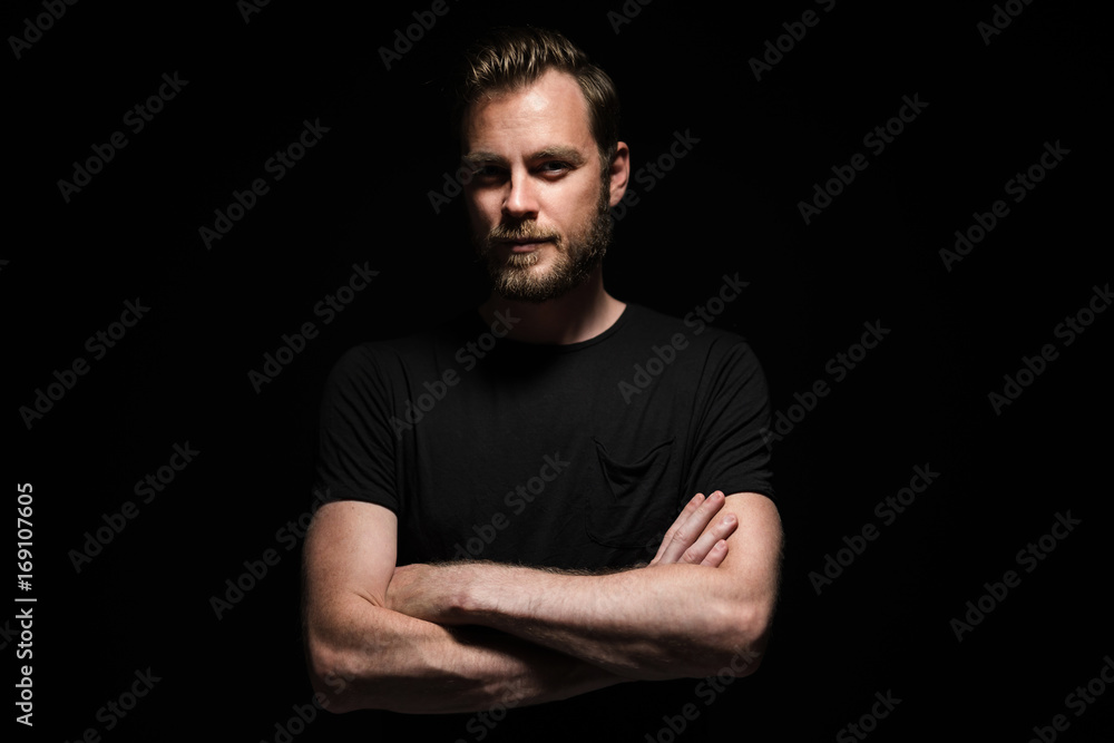 Portrait of a bearded man with an intense look standing in a dark room against a black background.