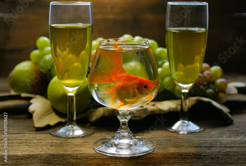 A fish in a glass against a background of grapes of wine glasses.