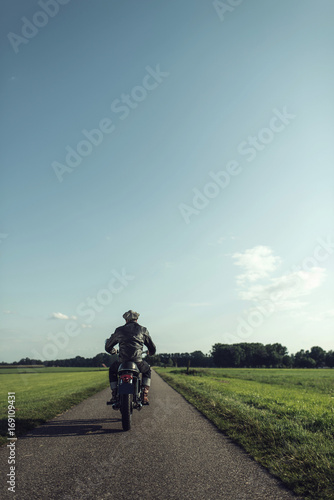 Rear view of motorcyclist riding vintage motorcycle on country road.