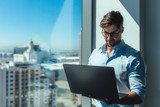 Business investor standing by a window holding a laptop.