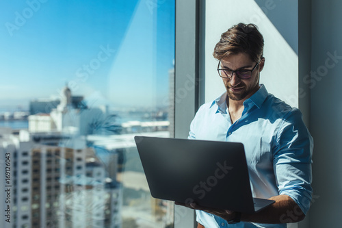 Business investor standing by a window holding a laptop.