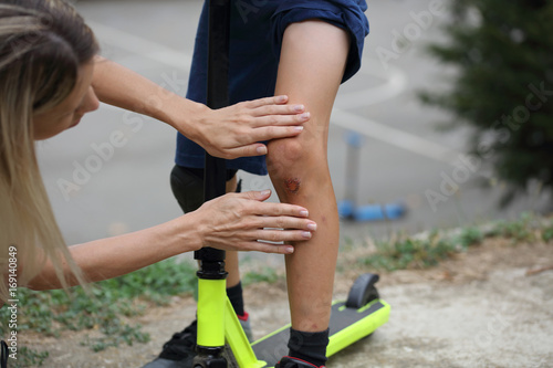 Mother treats her son skinned knee. Boy injured his leg during scooter riding. Kids safety ,using protective knee pads concept