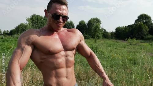 Muscular bodybuilder man standing, shirtless, showing chiselled torso, abs and pecs in green grass lawn in nature during a sunny summer day photo