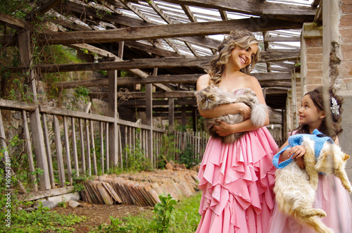 Beautiful woman and her cute little daughter looking each other and smiling wearing a princess dress, while they are hugging their cats, in outdoors