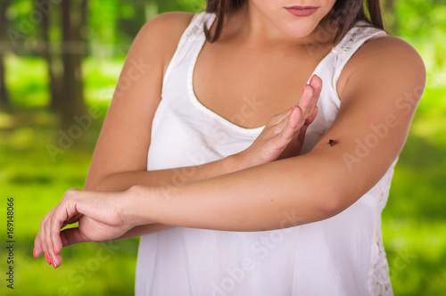 Fototapeta Naklejka Na Ścianę i Meble -  Close up of a young woman using her hand to kill a mosquito over her arm, in a blurred green background