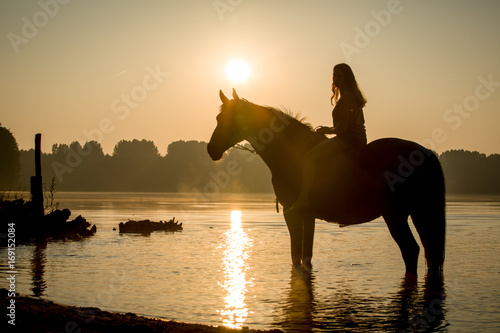 Reiterin mit Pferd im See im Sonnenaufgang