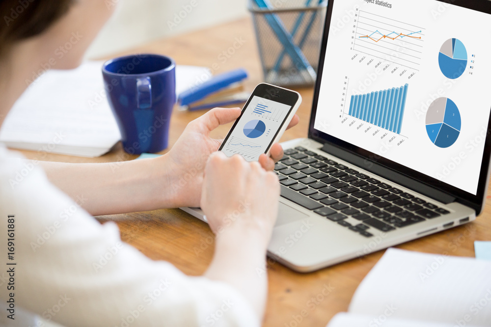 Businesswoman holding smartphone while working with laptop at home office desk, using cross platform responsive design apps for project analysis, analyzing cloud data statistics, close up rear view Businesswoman holding smartphone while working with laptop at home office desk, using cross platform responsive design apps for project analysis, analyzing cloud data statistics, close up rear view