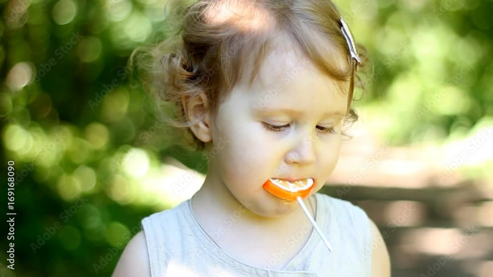 A child on the nature in the park sits and eats an orange candy. Lollipop A child on the nature in the park sits and eats an orange candy. Lollipop