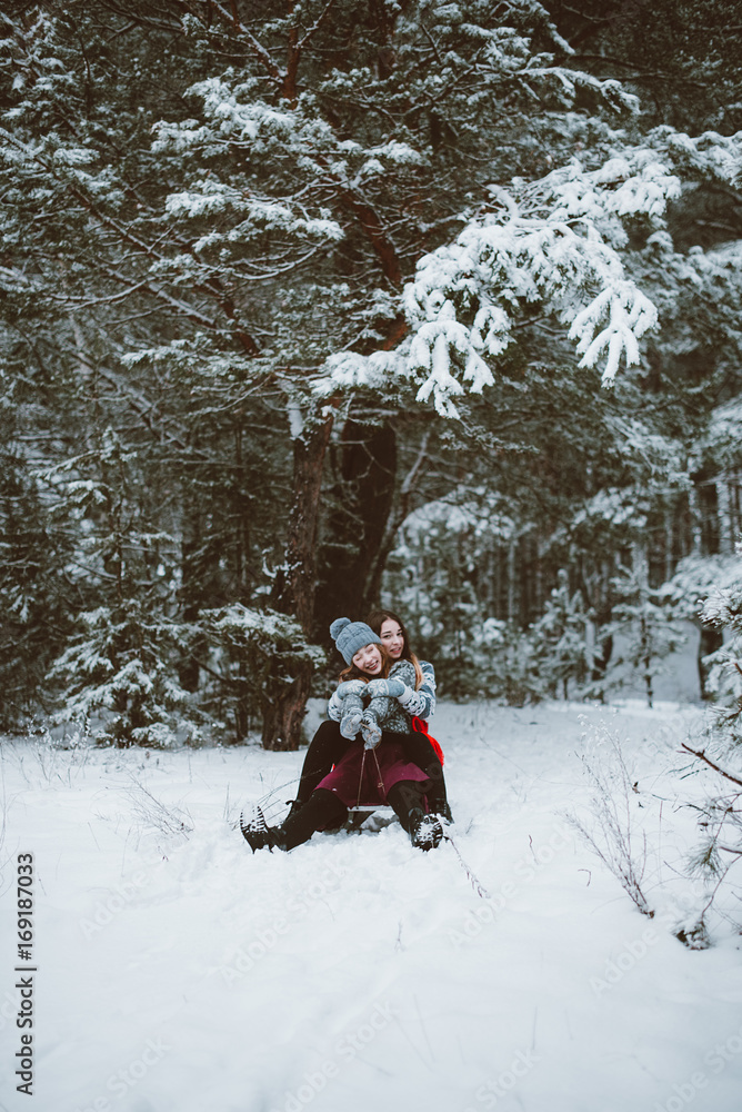 Close up fashion portrait of two sisters hugs and having fun, ride on sled in winter time forest, wearing sweaters and scarfs,best friends couple outdoors, snowy weather