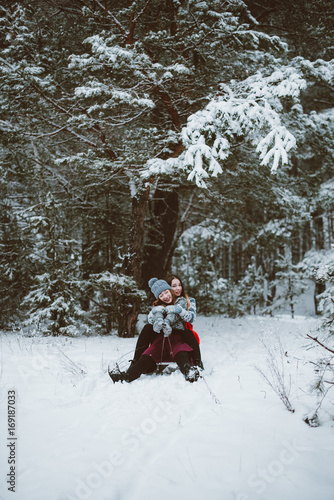 Close up fashion portrait of two sisters hugs and having fun, ride on sled in winter time forest, wearing sweaters and scarfs,best friends couple outdoors, snowy weather © dashamuller