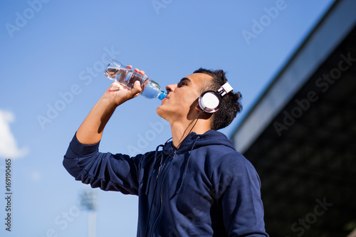 Good looking white male teenager wearing blue hoodie and headphones drinks water from bottle while looking at the sky