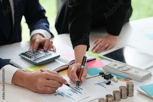 Business man and women are use pen pointing data together and have many coin, dollar cash, tablet, calculator decorate on white table, Closed up, Business and finance concept.