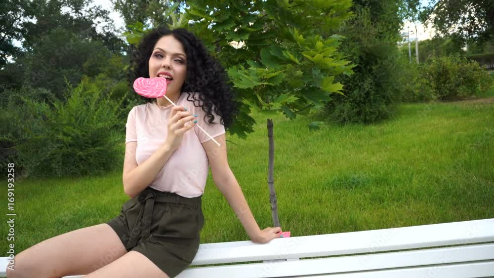 A teenage girl is eating lollipop, sitting on a bench in a city park. Girl waiting for a friend A teenage girl is eating lollipop, sitting on a bench in a city park. Girl waiting for a friend