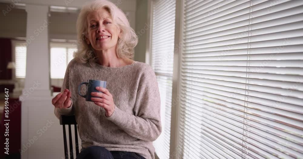 Cheerful elder woman holding coffee mug snapping her fingers smiling at camera. Senior Caucasian woman sitting by window dancing in her chair inside home. 4k Cheerful elder woman holding coffee mug snapping her fingers smiling at camera. Senior Caucasian woman sitting by window dancing in her chair inside home. 4k