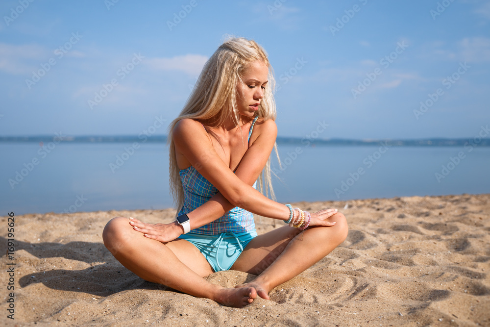 Blonde long haired girl in a blue suit is stretching and doing yoga on a lovely beach in sunlight of the rising sun