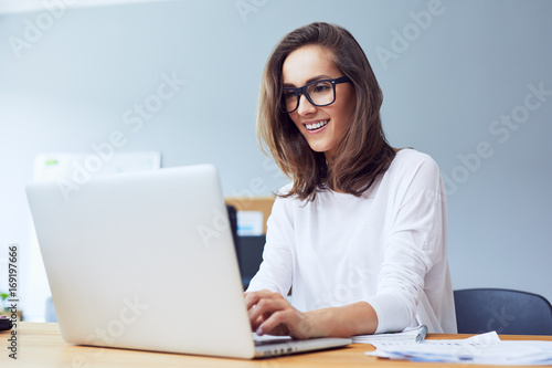 Front view of attractive lady working on laptop and smiling in modern home office