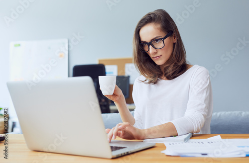 Focused young businesswoman holding coffee and using laptop in bright modern office