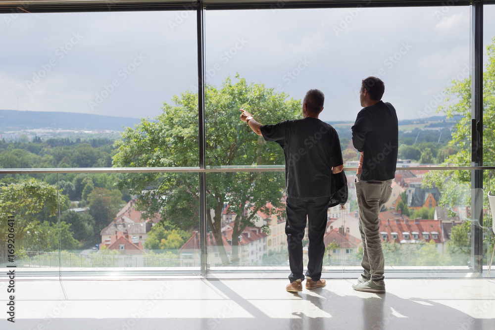 Two business man looking out of modern office window pointing with arm, from back Two business man looking out of modern office window pointing with arm, from back