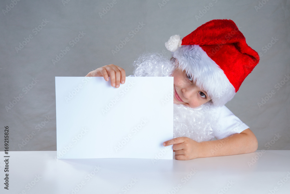 Little Santa holding a white envelope, smiling. Sitting behind a white table looking at the camera. Close-up. Gray background. Little Santa holding a white envelope, smiling. Sitting behind a white table looking at the camera. Close-up. Gray background.