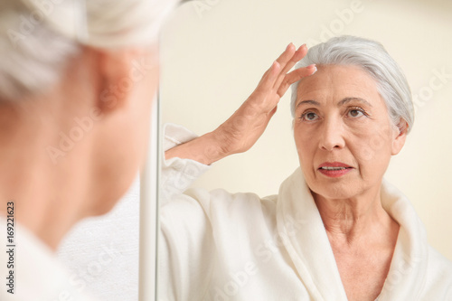 Beautiful elderly woman applying face cream in front of mirror at home