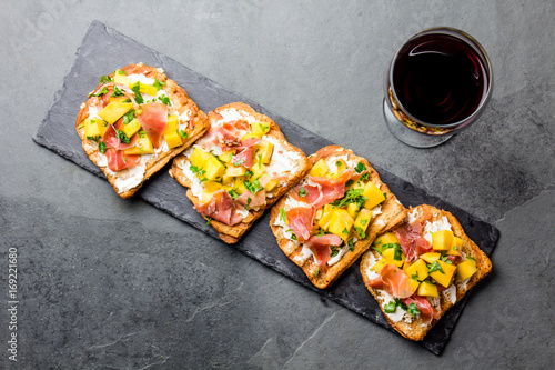 Toasts with cream cheese, ham jamon serrano and mango served on black slate board with red wine, top view, gray slate background