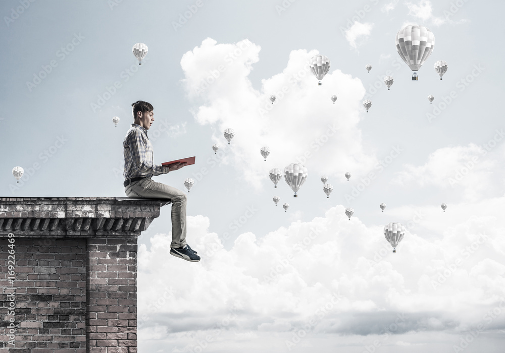 Man on roof edge reading book and aerostats flying in sky Man on roof edge reading book and aerostats flying in sky