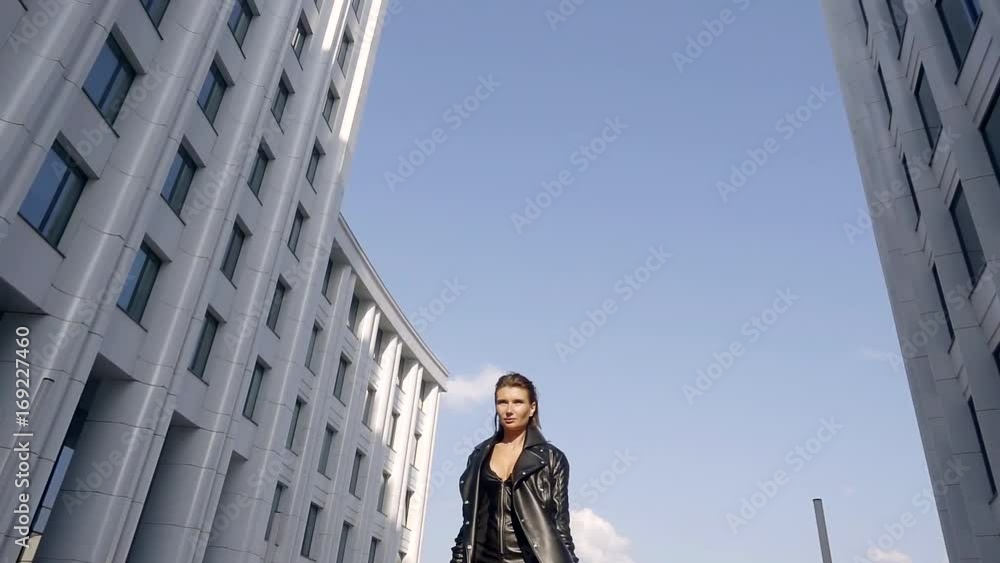 slow motion. 500 frames per second. fashion stylish slender young girl dressed in a jumpsuit and Cape eco leather is daytime on the street on the background of clear blue sky and the modern city slow motion. 500 frames per second. fashion stylish slender young girl dressed in a jumpsuit and Cape eco leather is daytime on the street on the background of clear blue sky and the modern city