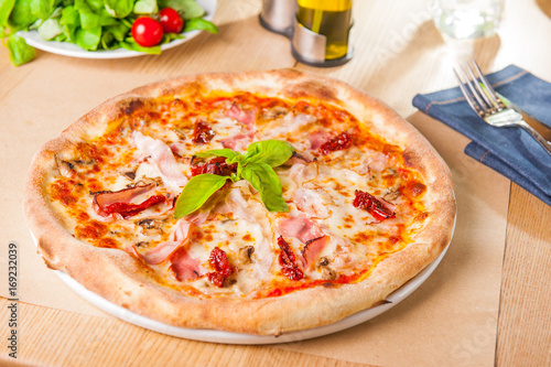 Italian fast food. Delicious hot Meat Pizza with bacon and sun dried tomatoes decorated with bazil leaves and served on wooden platter with ingredients, close up view. Menu photo. Selective focus